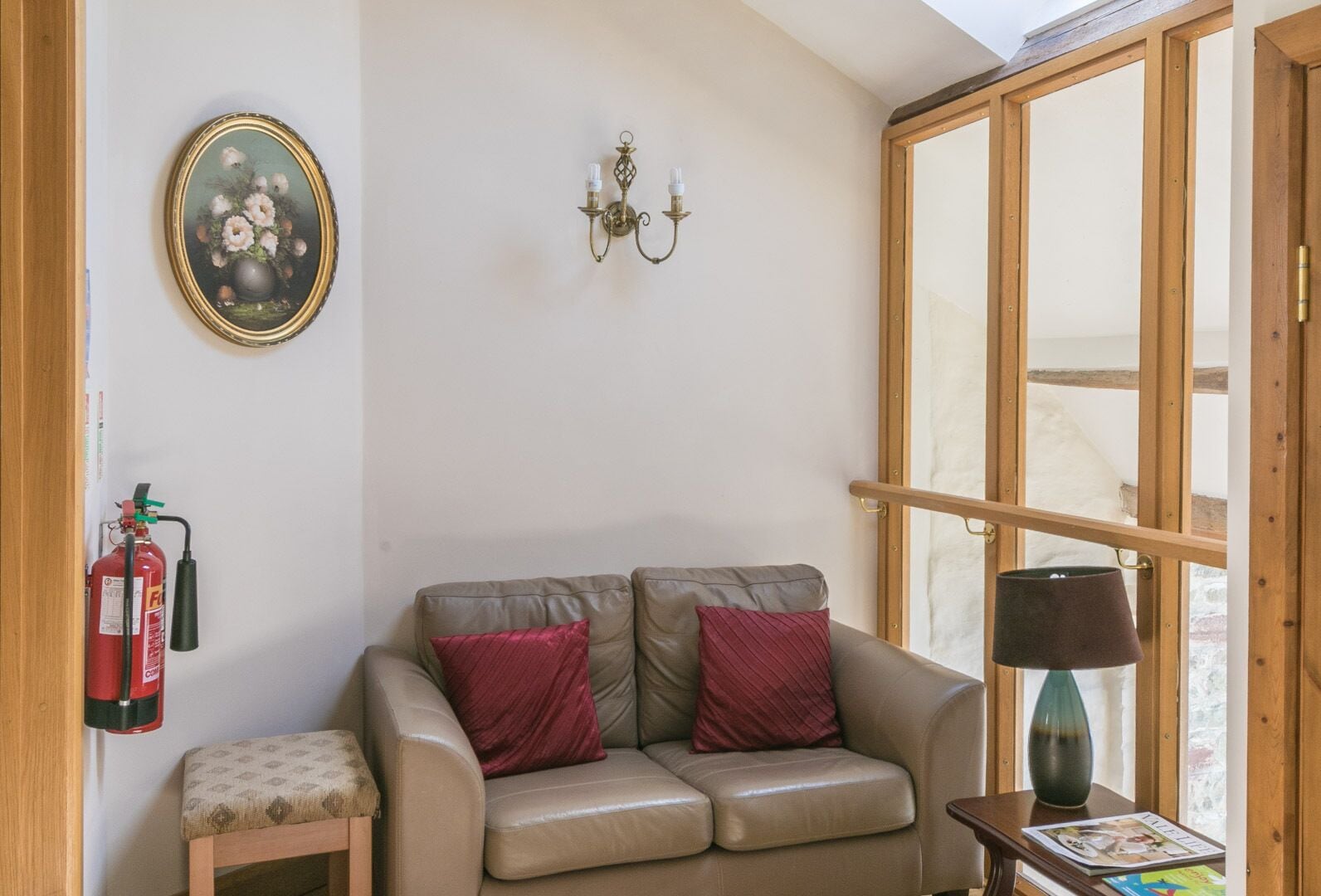 A cozy living room with a beige leather sofa, two red cushions, a small side table with a lamp, a framed floral painting, a wall-mounted light fixture, and a fire extinguisher.