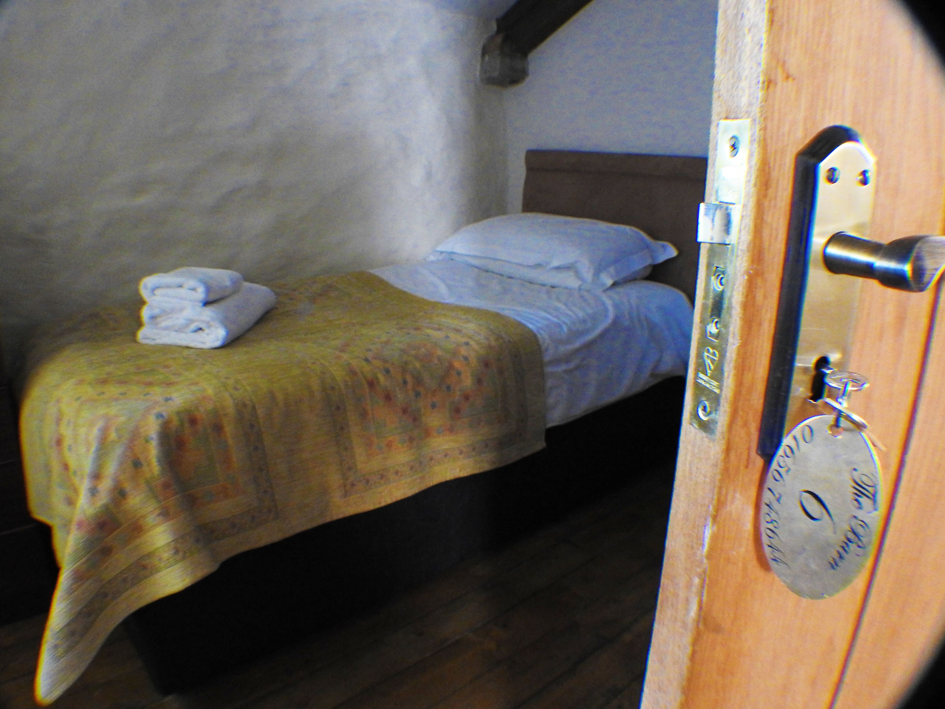 Una cama individual con una colcha amarilla, almohadas blancas y una toalla doblada en la cabecera. Se ve una puerta de madera con manija de metal y una placa de latón.