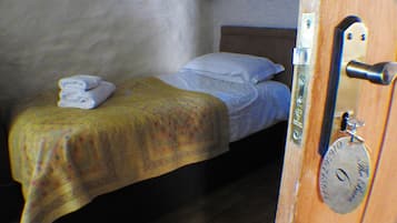Una cama individual con una colcha amarilla, almohadas blancas y una toalla doblada en la cabecera. Se ve una puerta de madera con manija de metal y una placa de latón.
