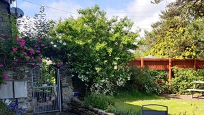 A patio with stone tiles, a metal table and chairs, and a garden with various plants and a wooden fence. - The Barn B&B (Port Talbot)