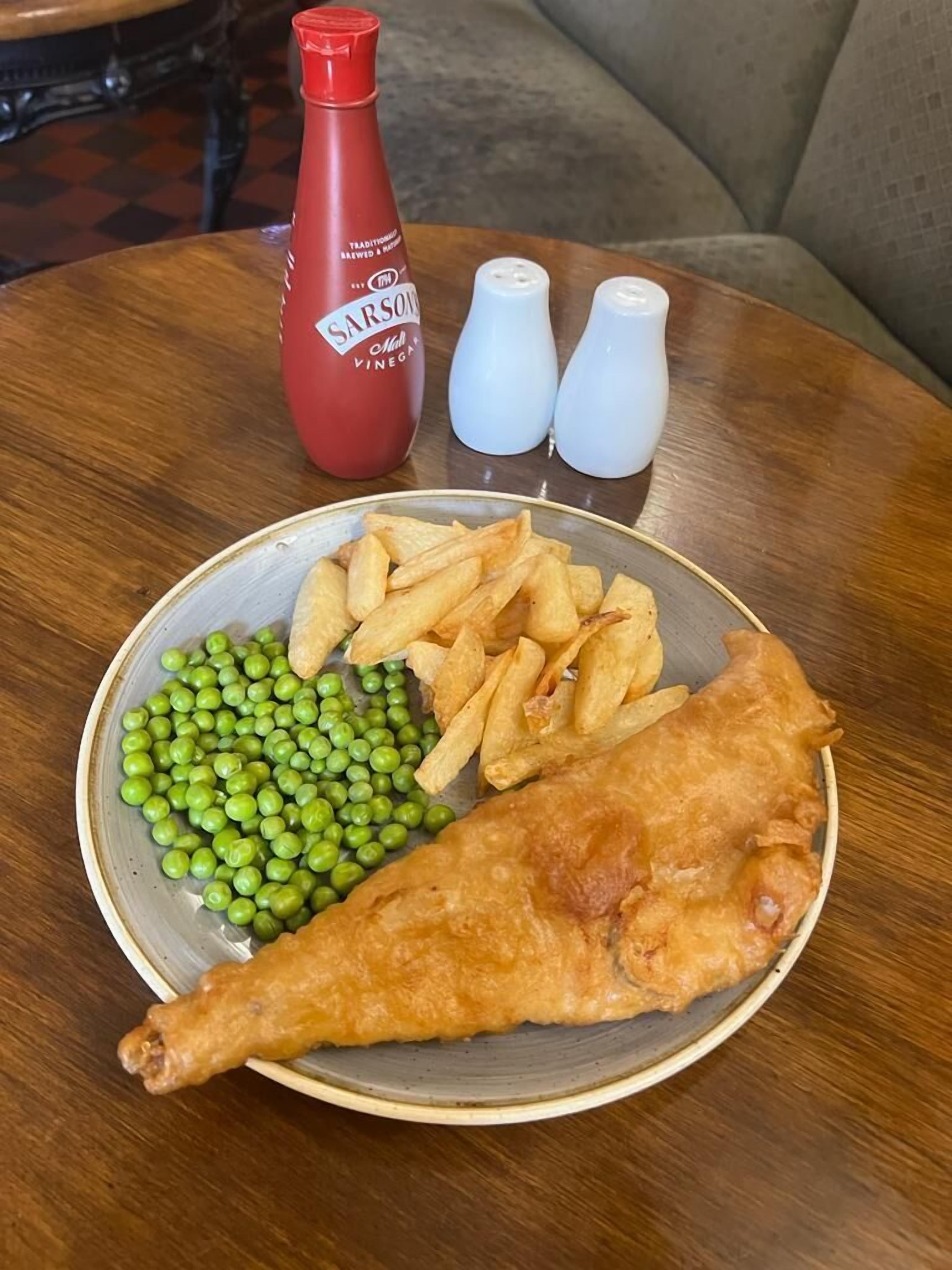 Un plato con pescado frito, guisantes y papas fritas sobre una mesa redonda de madera.