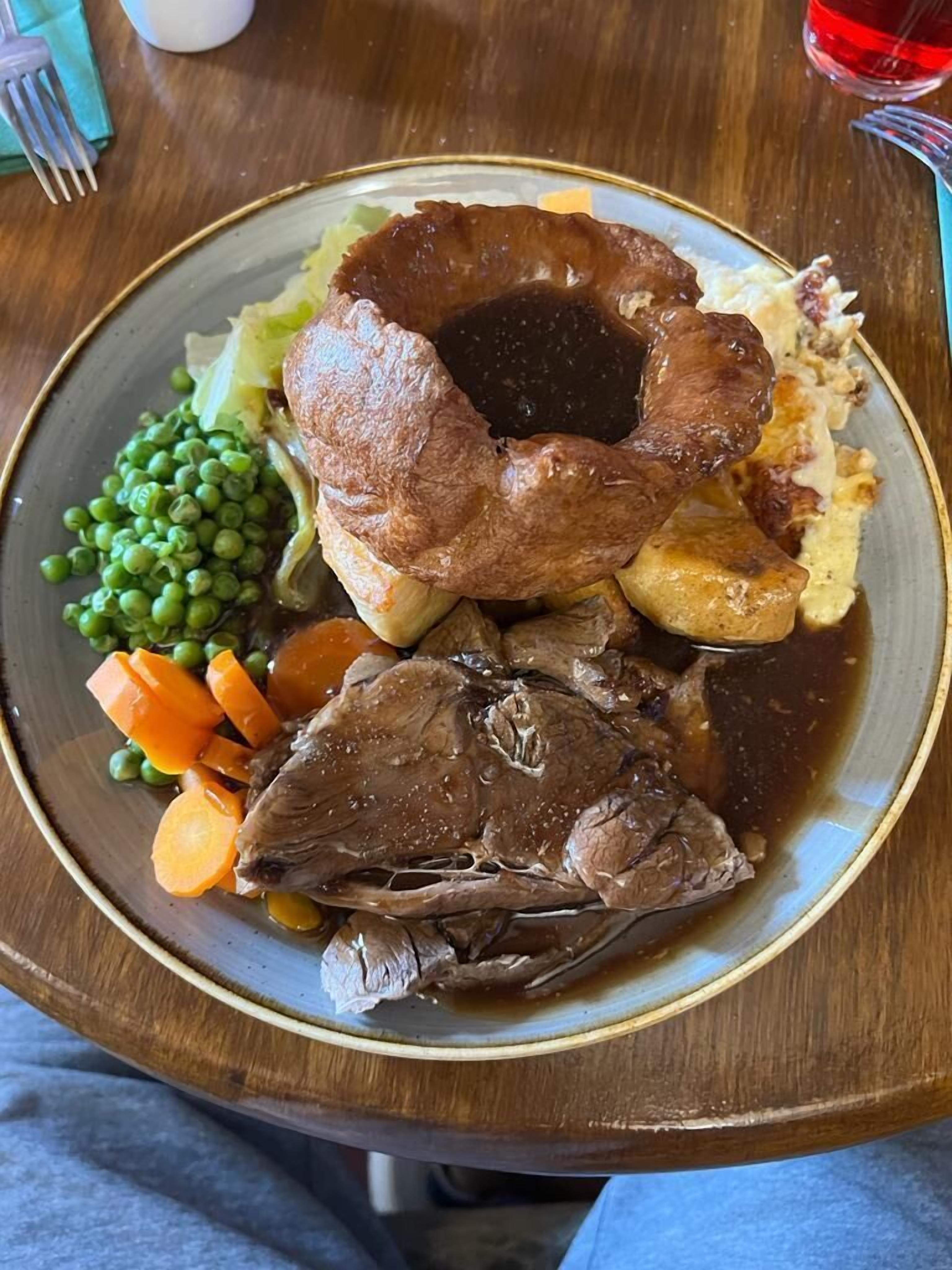 A plate of roast beef, Yorkshire pudding, peas, carrots, and mashed potatoes.