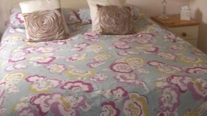 Una cama con cubrecama estampado con motivos florales, dos almohadas con diseño en espiral, una mesita de noche con lámpara y un buró con una caja de pañuelos.