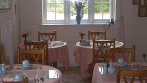Un comedor con cuatro mesas, cada una con mantel rosa y adornada con teteras y tazas. Hay dos sillas en cada mesa. Se ve una ventana con vistas a la vegetación al fondo.