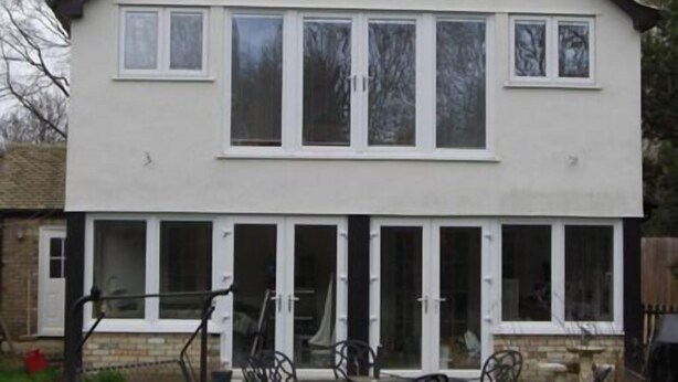 Una casa de dos pisos con un amplio ventanal frontal y un patio con mobiliario exterior.
