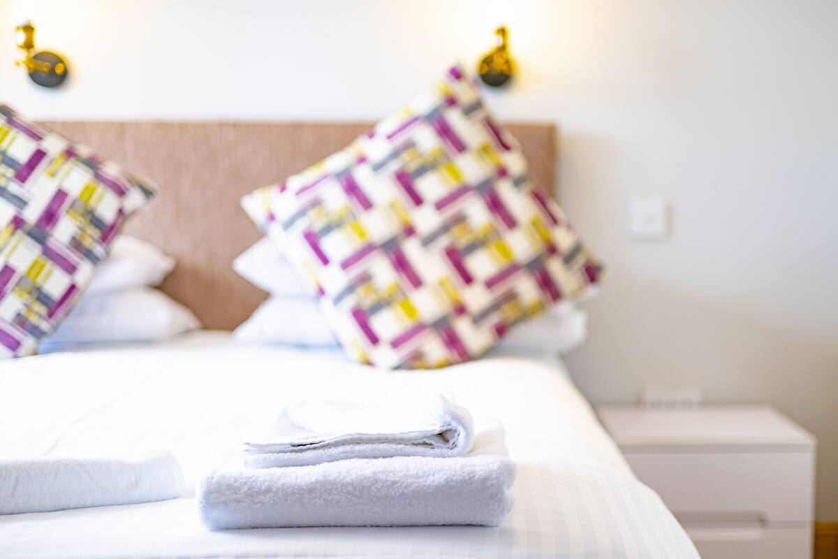 A neatly made bed with folded white towels and patterned pillows, a bedside table, and wall-mounted lamps.