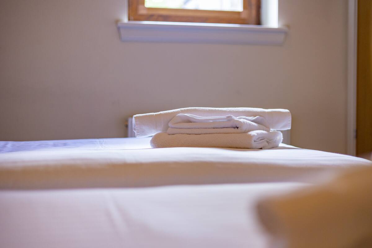 A neatly made bed with a folded towel on it, a window above the bed, and a white wall.
