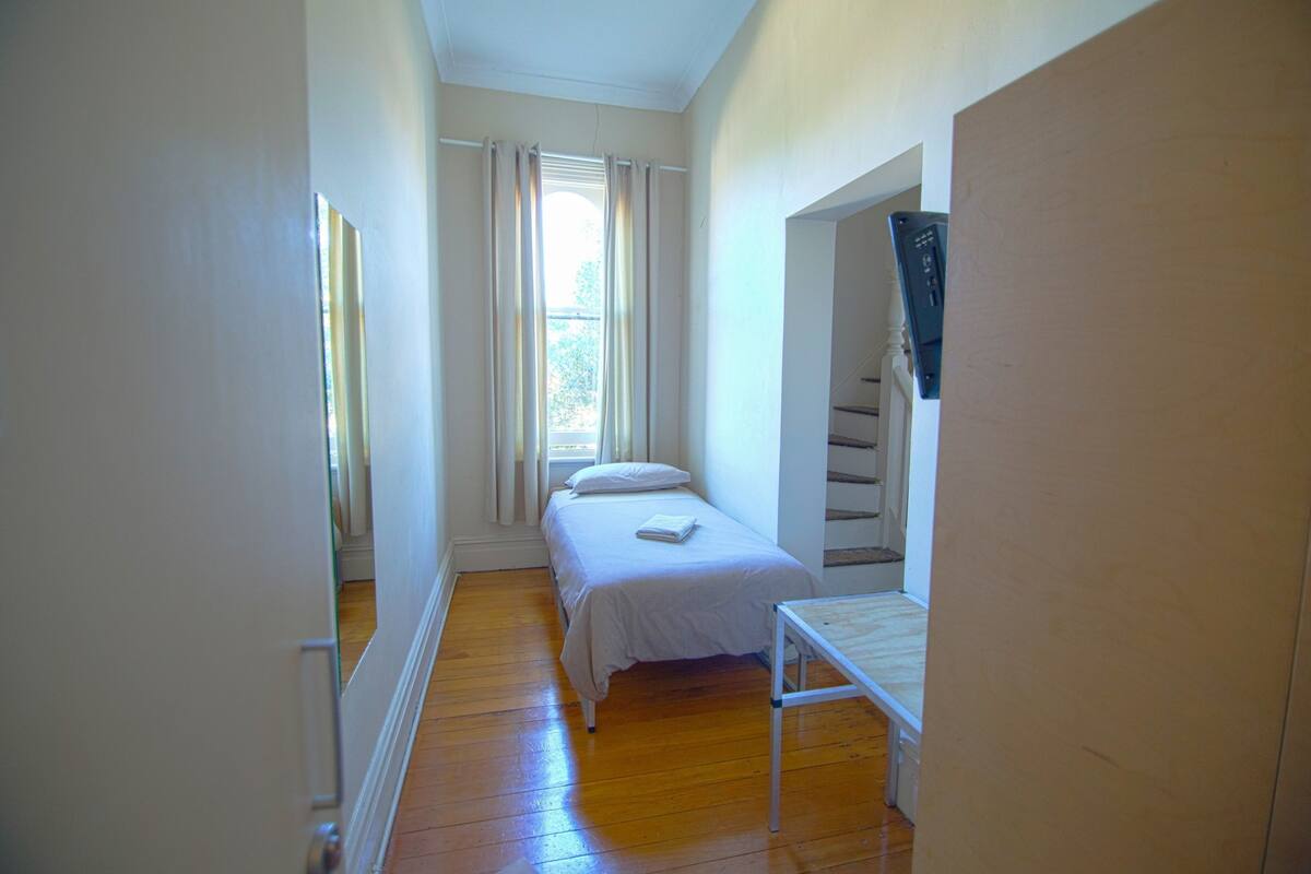 A narrow room with a single bed, a small table, and a window with curtains.