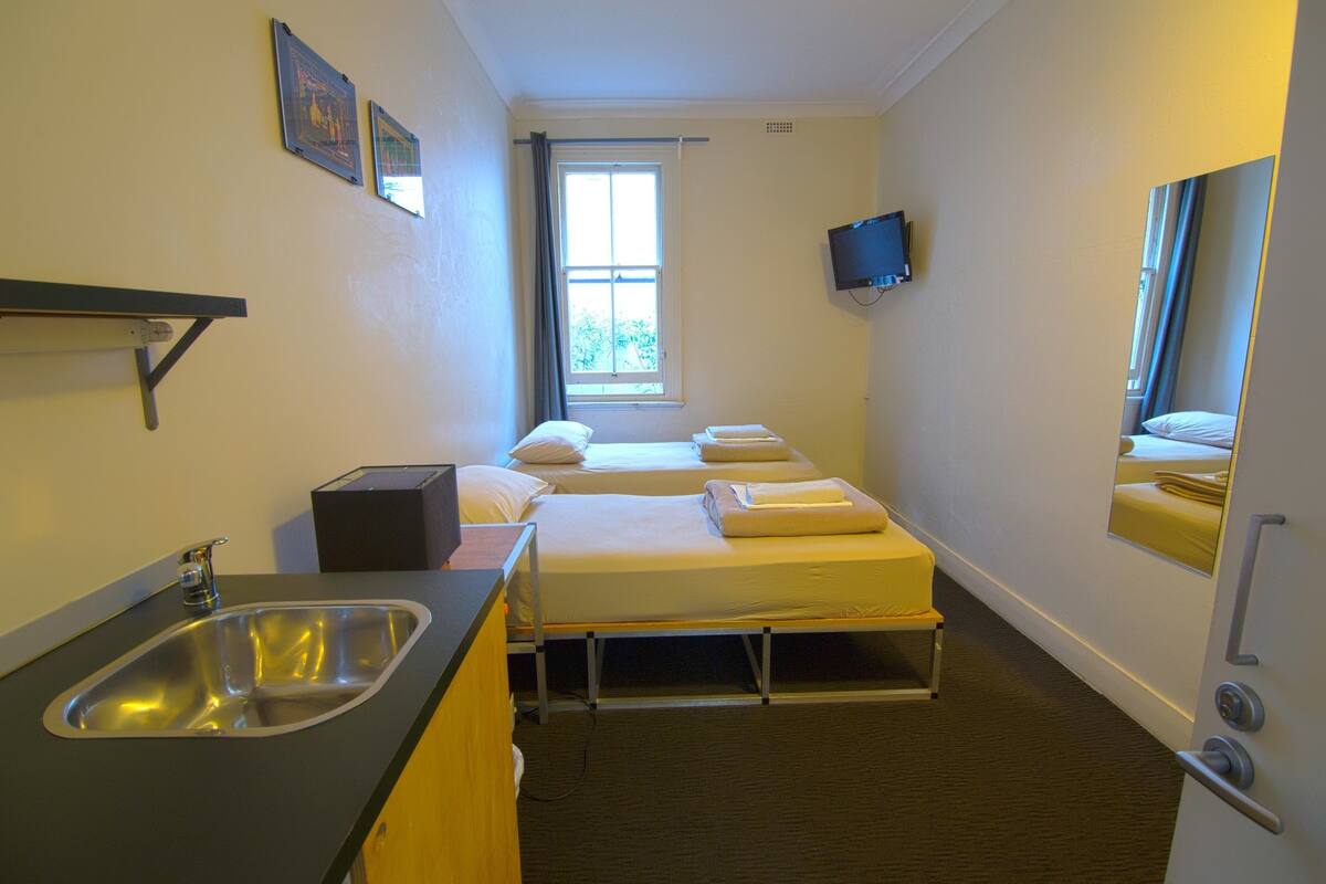 A compact hotel room with a single bed, a small sink, a television mounted on the wall, and a window providing natural light.