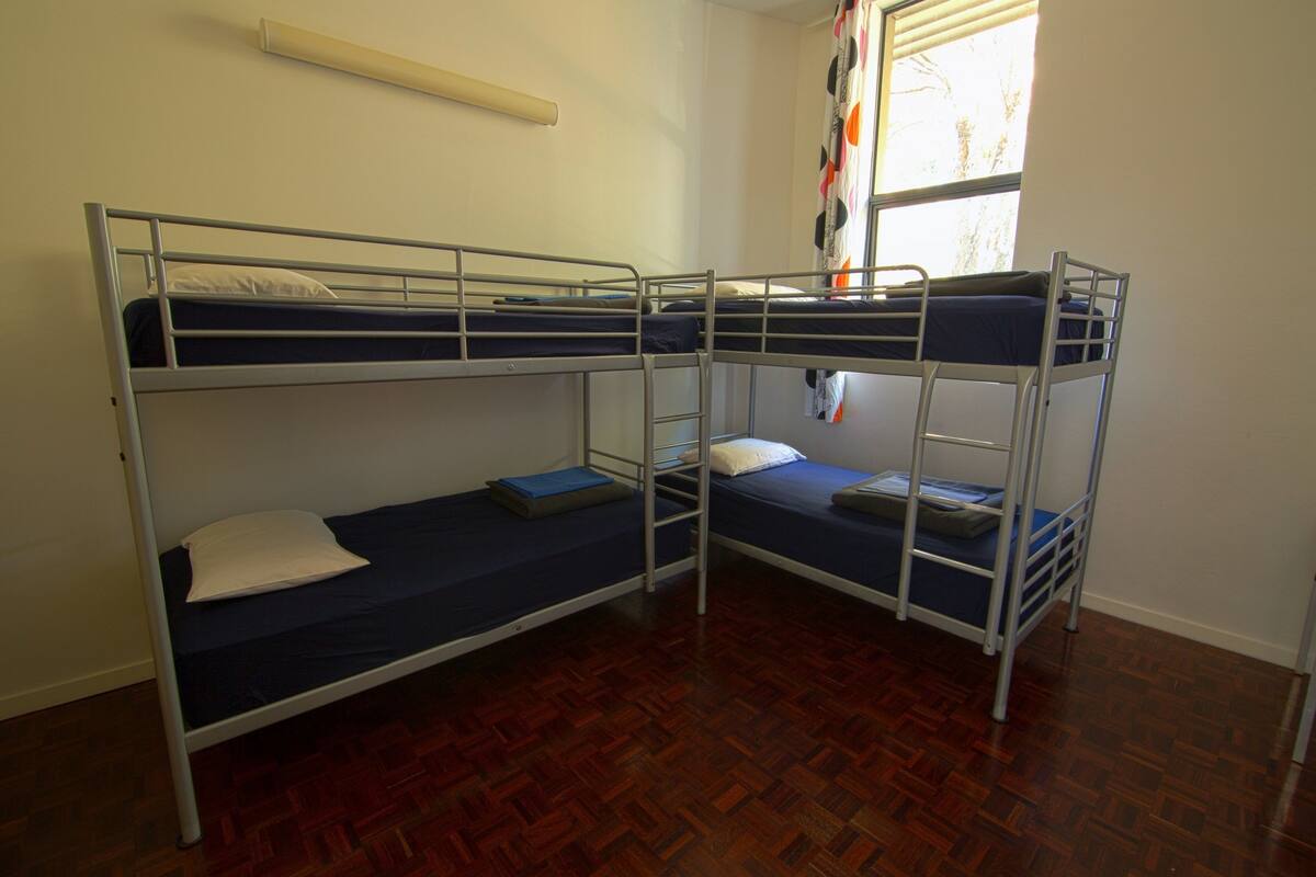 A room with three bunk beds, wooden parquet flooring, a window with curtains, and a wall-mounted shelf.