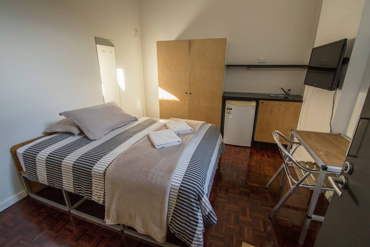 A compact room with a bed, a small kitchenette, and a TV mounted on the wall.