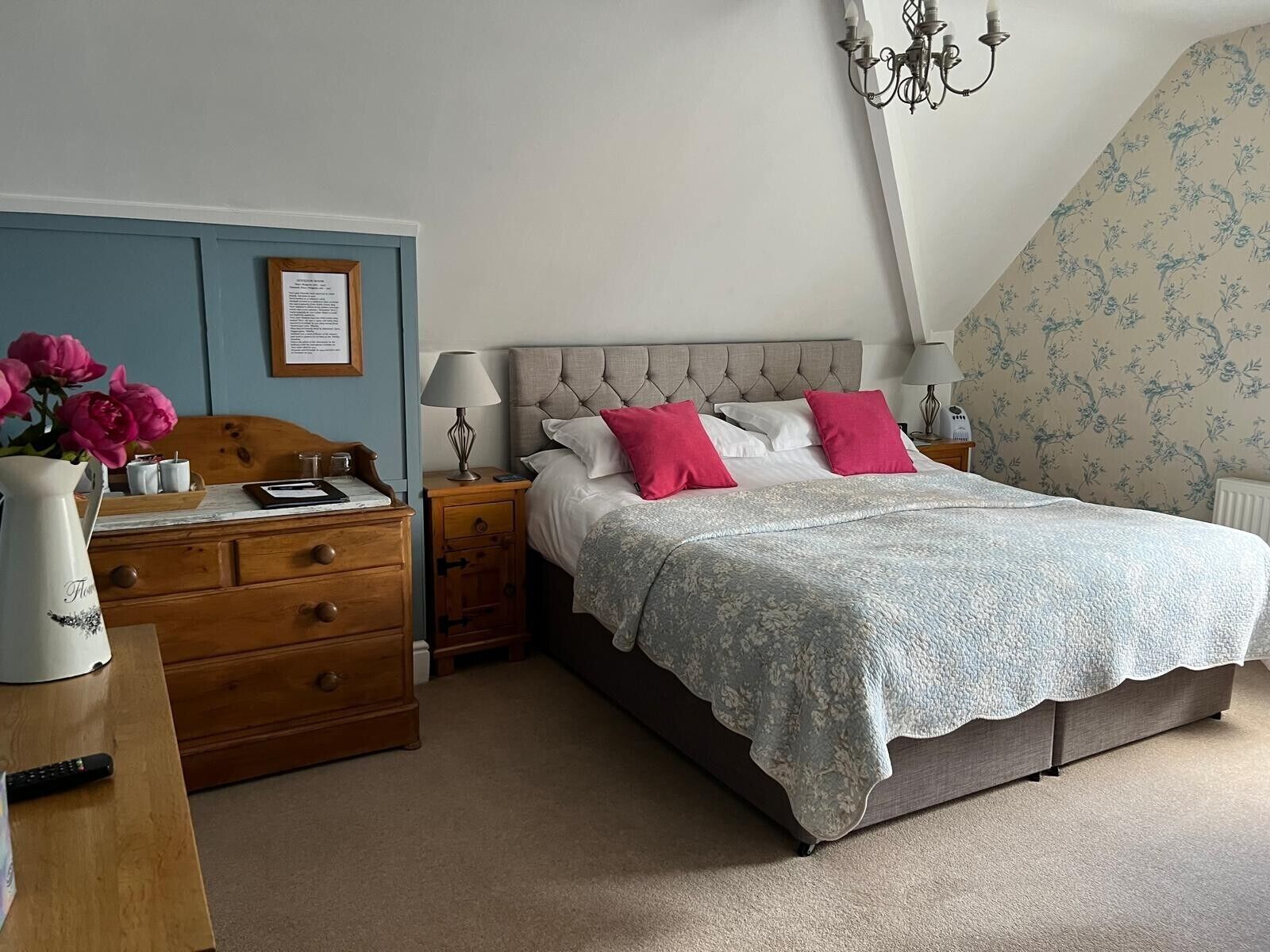 Quarto com cama, mesas de cabeceira, cômoda, um vaso com flores, um lustre e um documento emoldurado na parede.