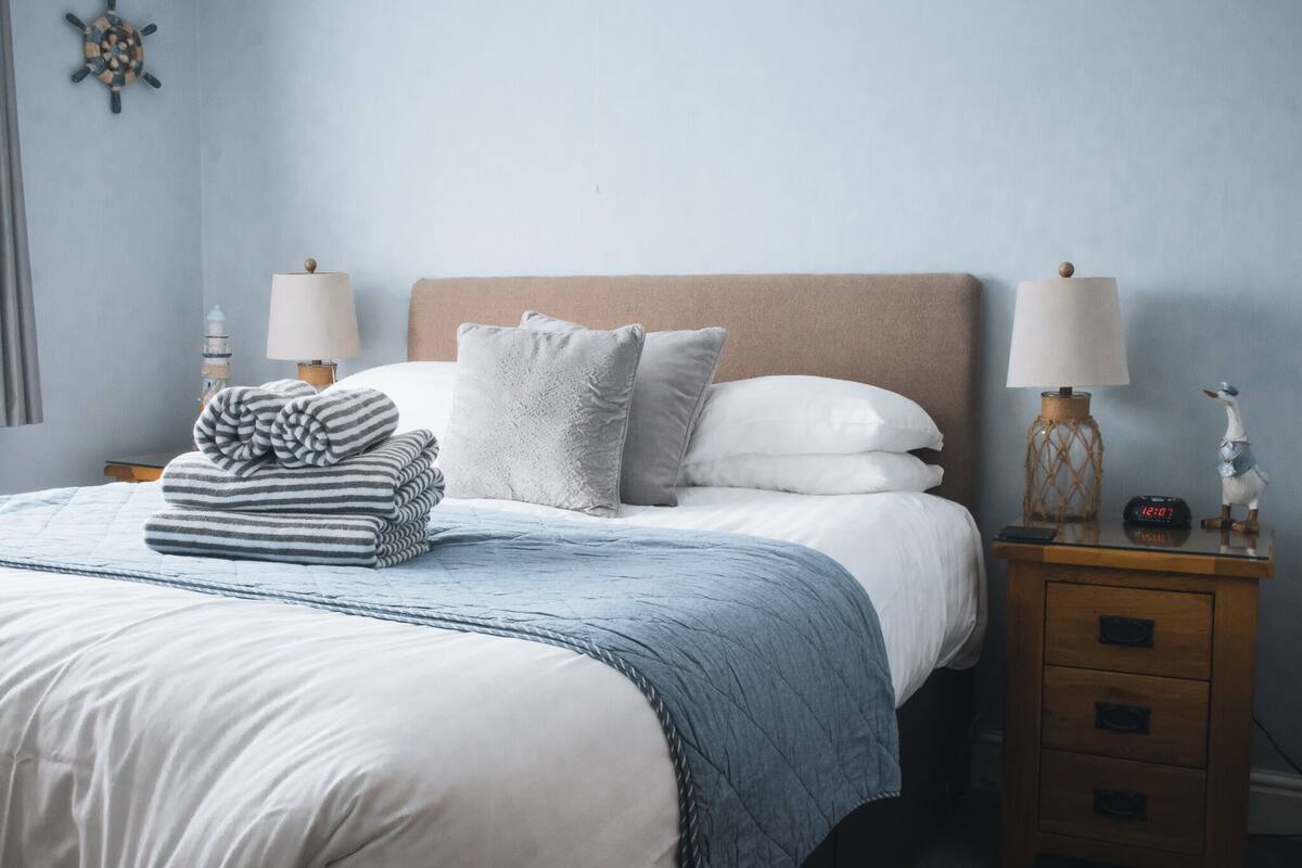 Una cama bien hecha con un edredón a rayas, dos almohadas y una colcha azul. Hay dos mesitas de noche con lámparas y una pequeña figurita en una de ellas.
