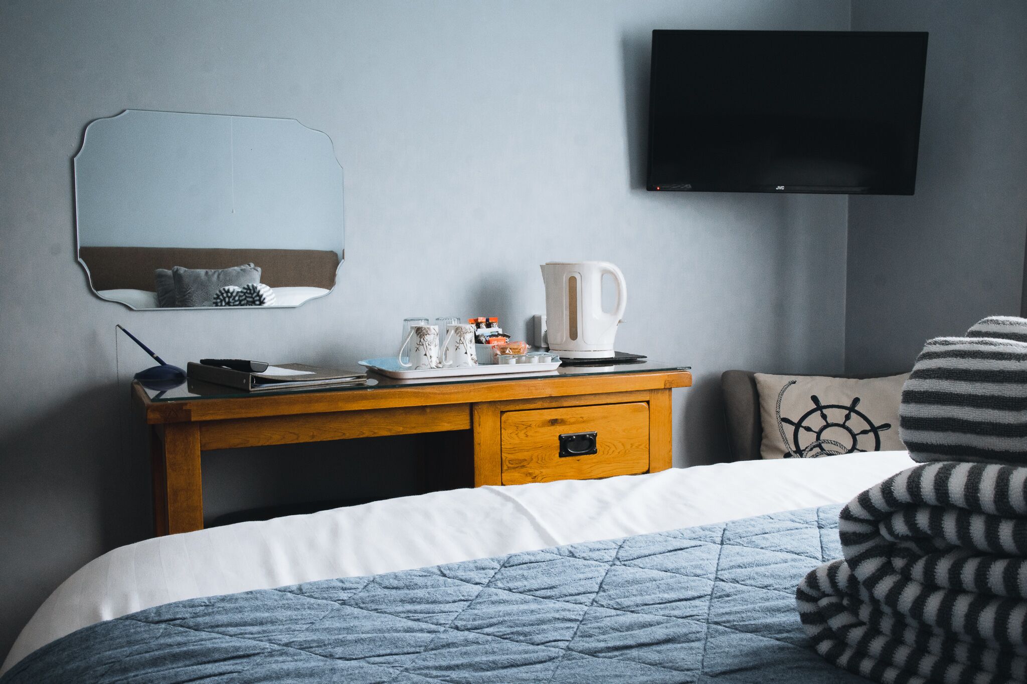 A neatly arranged hotel room with a bed, a bedside table with a kettle, a mirror, and a flat-screen TV on the wall.