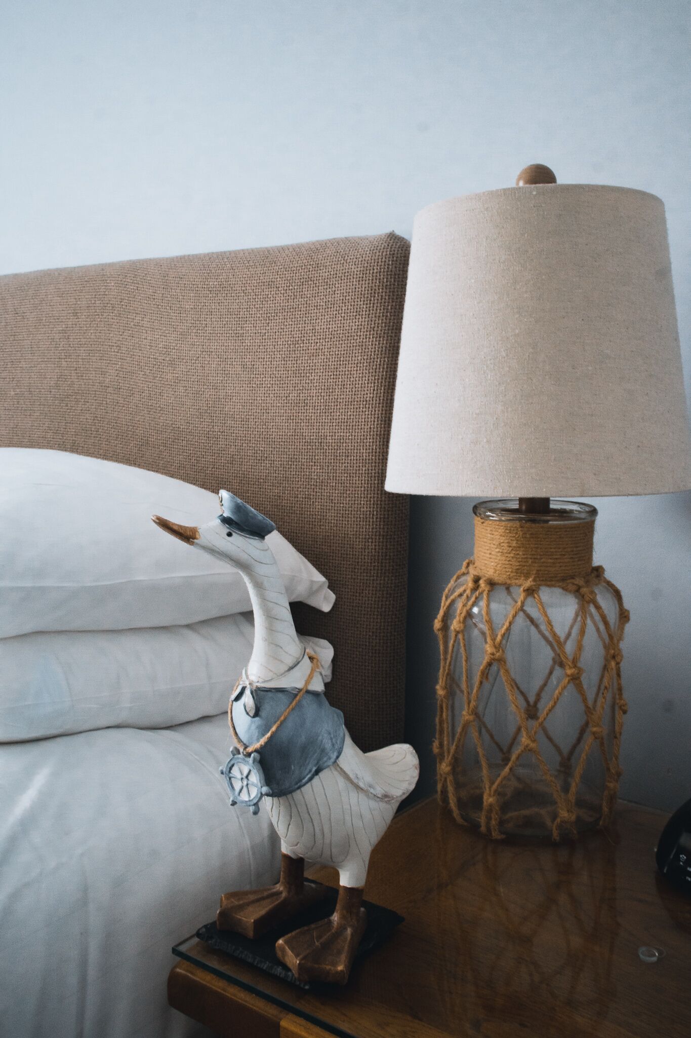 Een bed met witte lakens, een decoratief houten nachtkastje, een lamp met gevlochten voet en een beeldje van een vogel met hoed en sjaal.