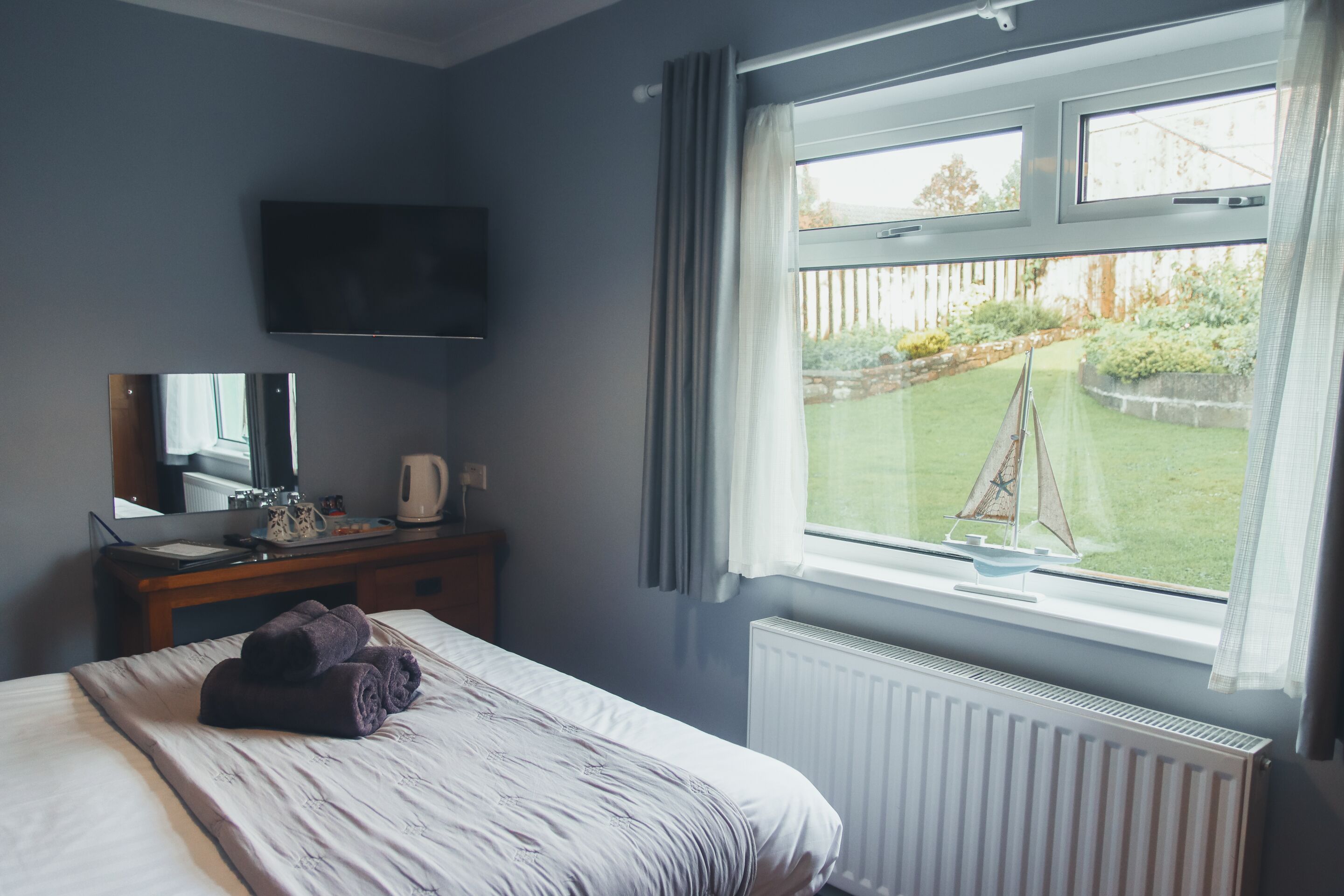 Un dormitorio con cama, escritorio con televisor de pantalla plana, ventana con cortinas y vista a un jardín.
