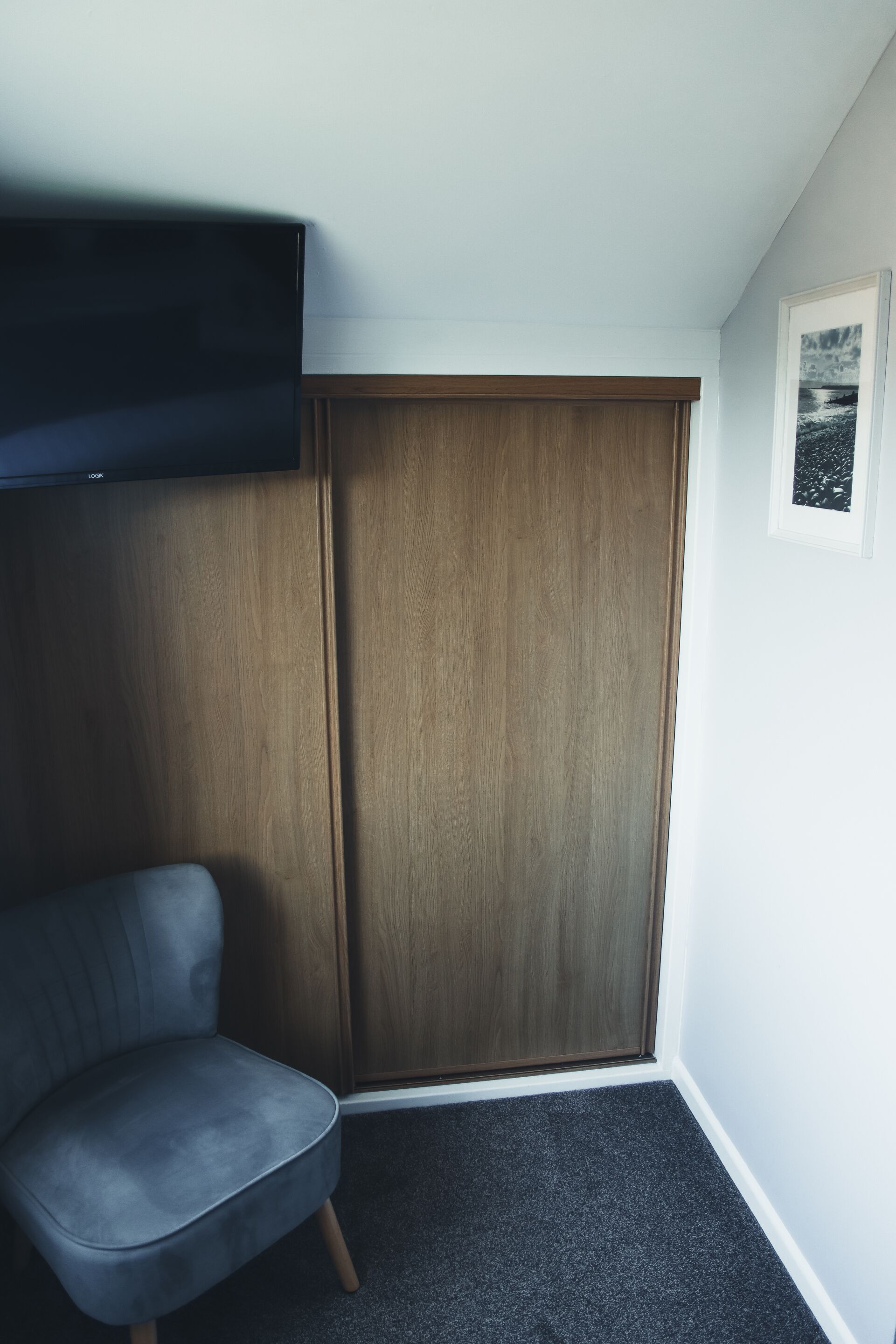 A corner room with a grey upholstered chair, a wooden wardrobe, and a framed picture on the wall.