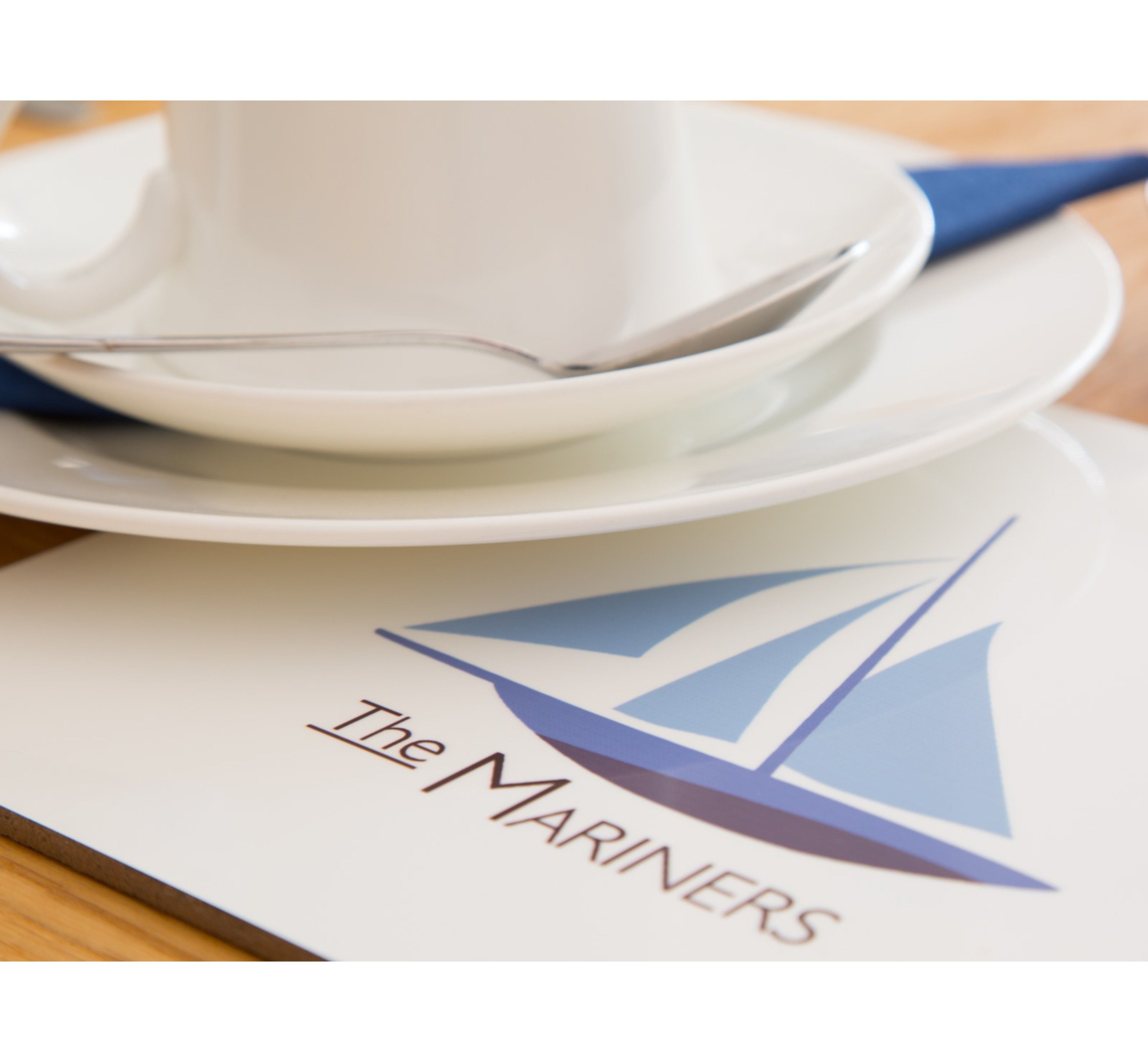 Uma mesa posta com xícara e pires brancos, uma colher e um logotipo com um veleiro.