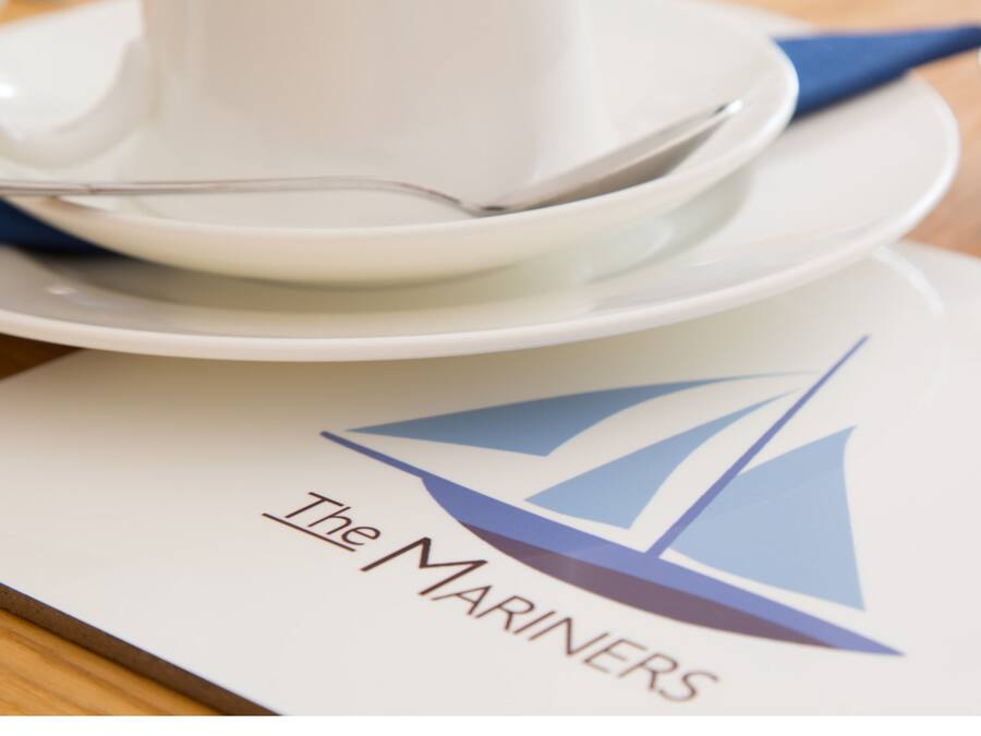 Uma mesa posta com xícara e pires brancos, uma colher e um logotipo com um veleiro.