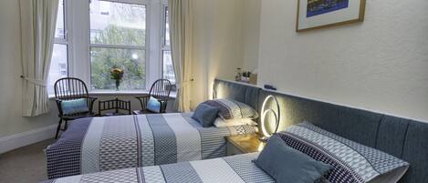 A hotel room with two beds, a window with curtains, a chair, a bedside table with a lamp, and a framed picture on the wall.