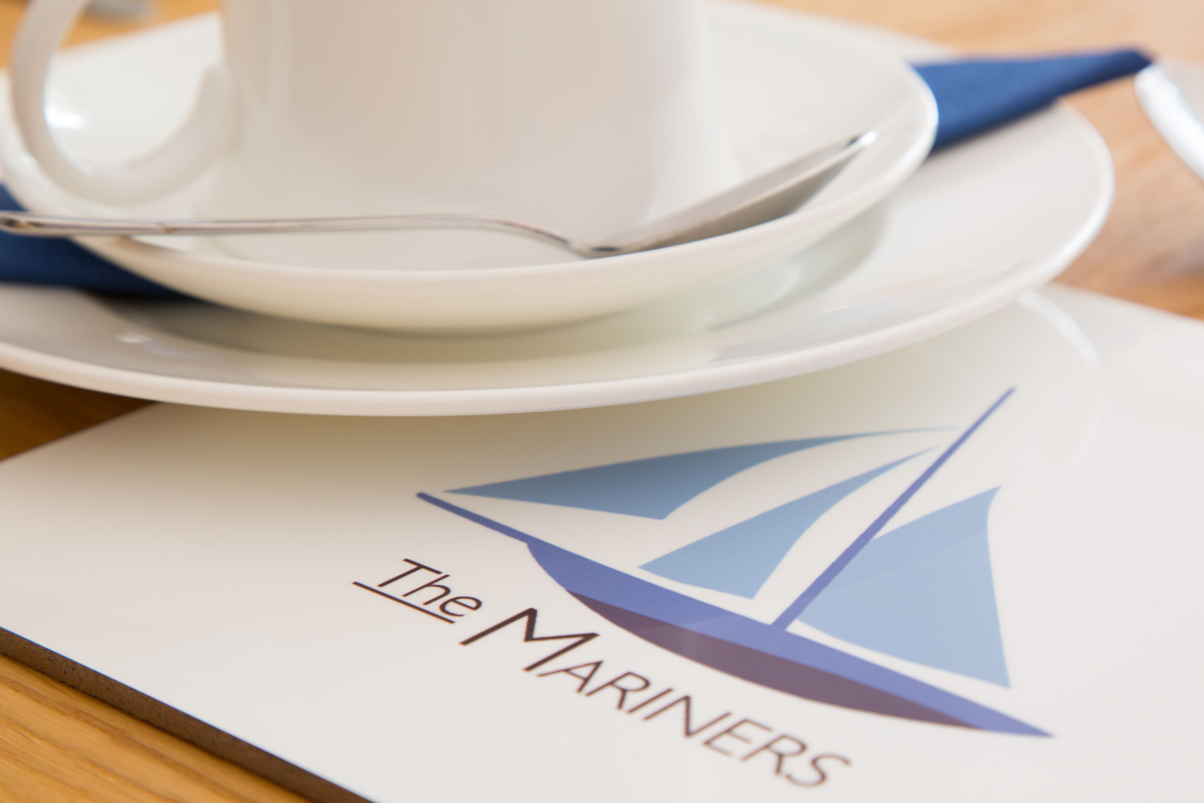 A table setting with a white cup, saucer, and spoon, and a menu with a sailboat logo and the text "The Mariners".
