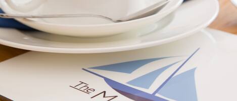 A table setting with a white cup, saucer, and spoon, and a menu with a sailboat logo and the text "The Mariners".