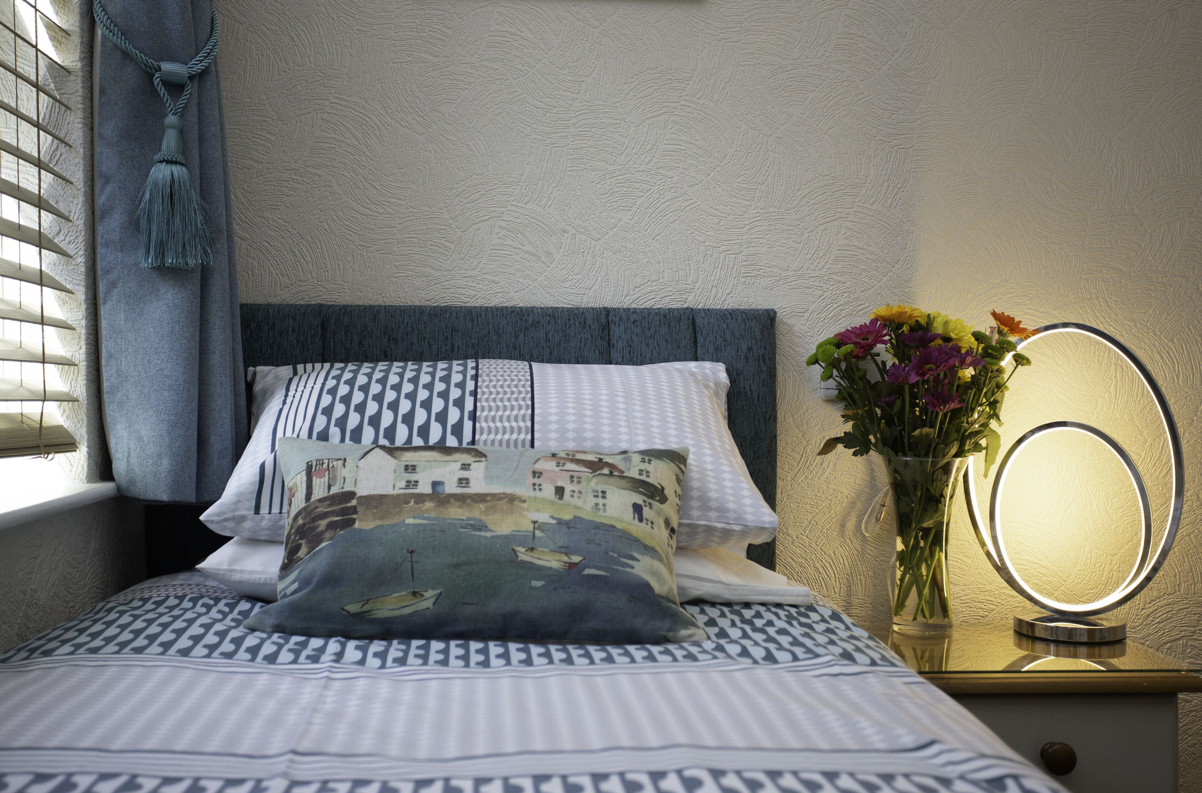 A neatly made bed with patterned bedding, a bedside mirror with a light, and a vase with flowers on a nightstand.