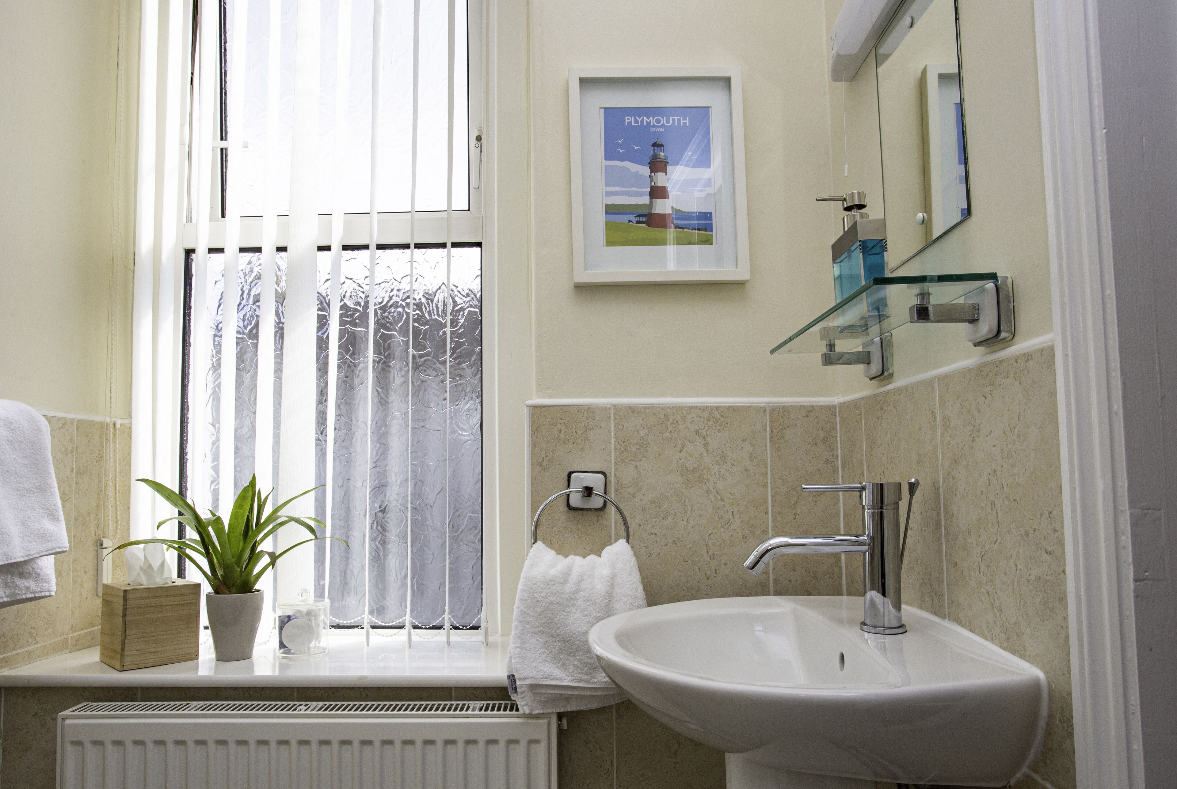 Een badkamer met een wit wastafelmeubel, een potplant, een lijst met een foto van een vuurtoren en een raam met gezandstraald glas.