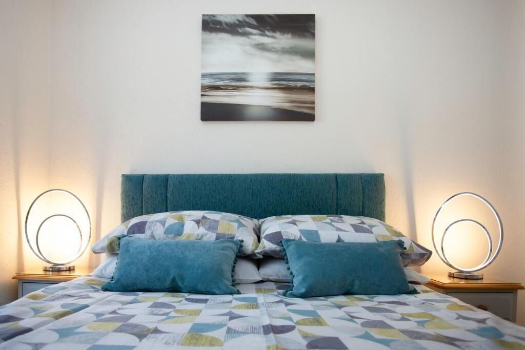 A bed with a geometric patterned bedspread, a teal upholstered headboard, and two bedside tables with illuminated mirrors.