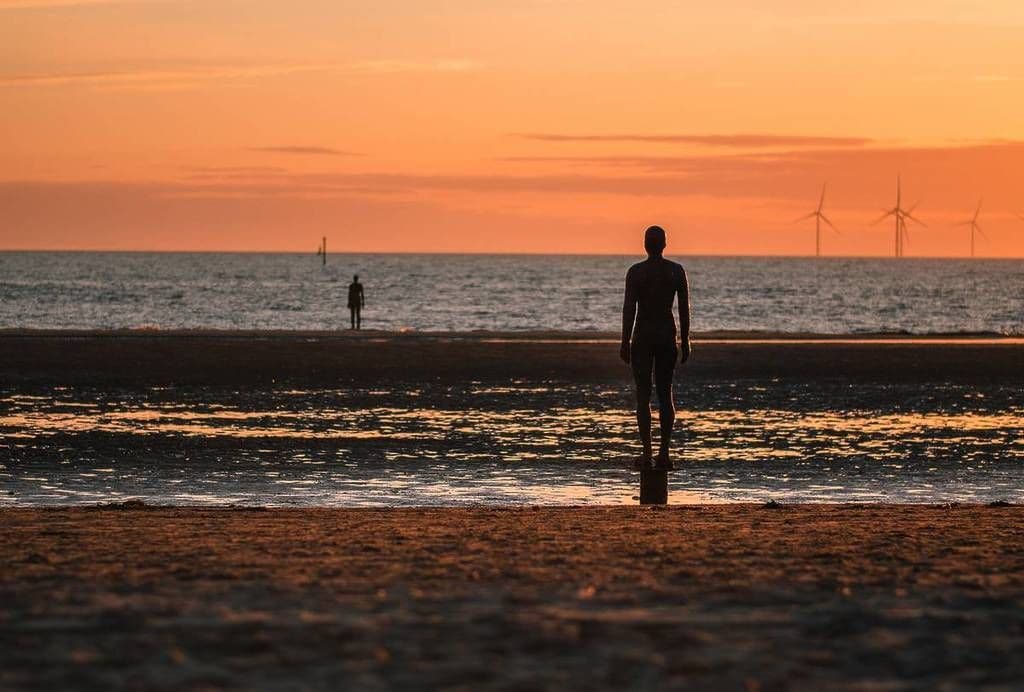 A beach scene with a person standing facing the ocean, with wind turbines in the distance.