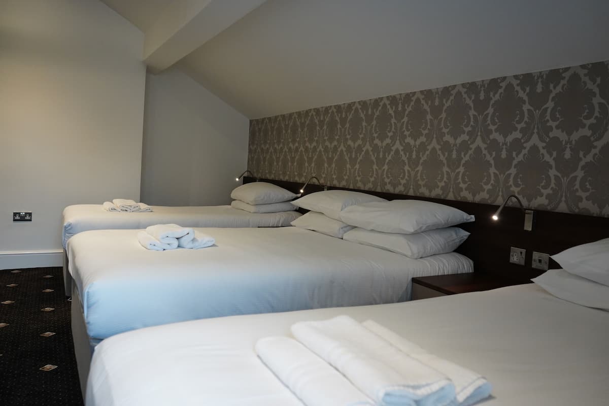 A hotel room with three beds, each with white linens and pillows, a patterned wallpaper on the wall, and a small bedside table with a lamp.