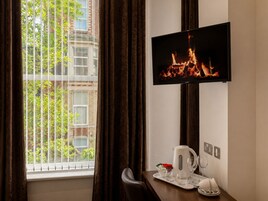 Una camera d'albergo con una televisione che mostra un caminetto, una finestra con tende e un tavolino con bollitore e bicchieri.