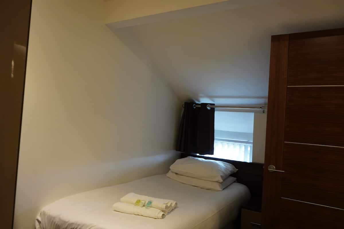 A small, single-bed room with a wooden door, a window with curtains, and a neatly made bed with a towel.