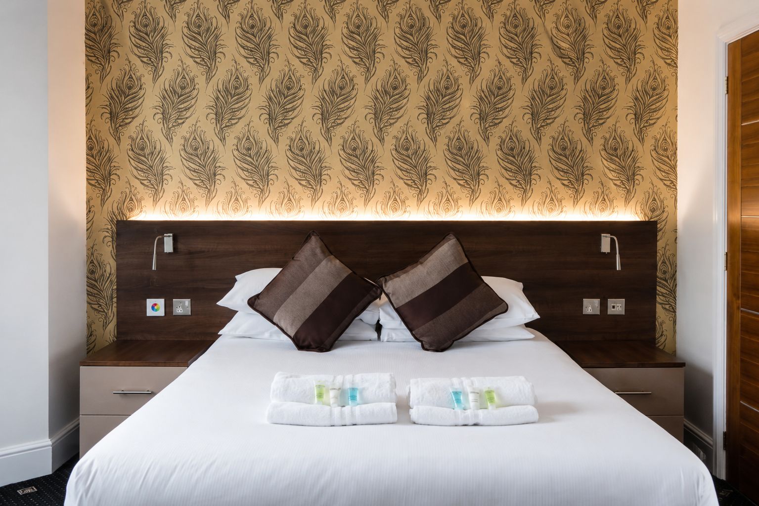 Habitación de hotel con cama, dos mesitas de noche, cabecero con luz y papel tapiz estampado.