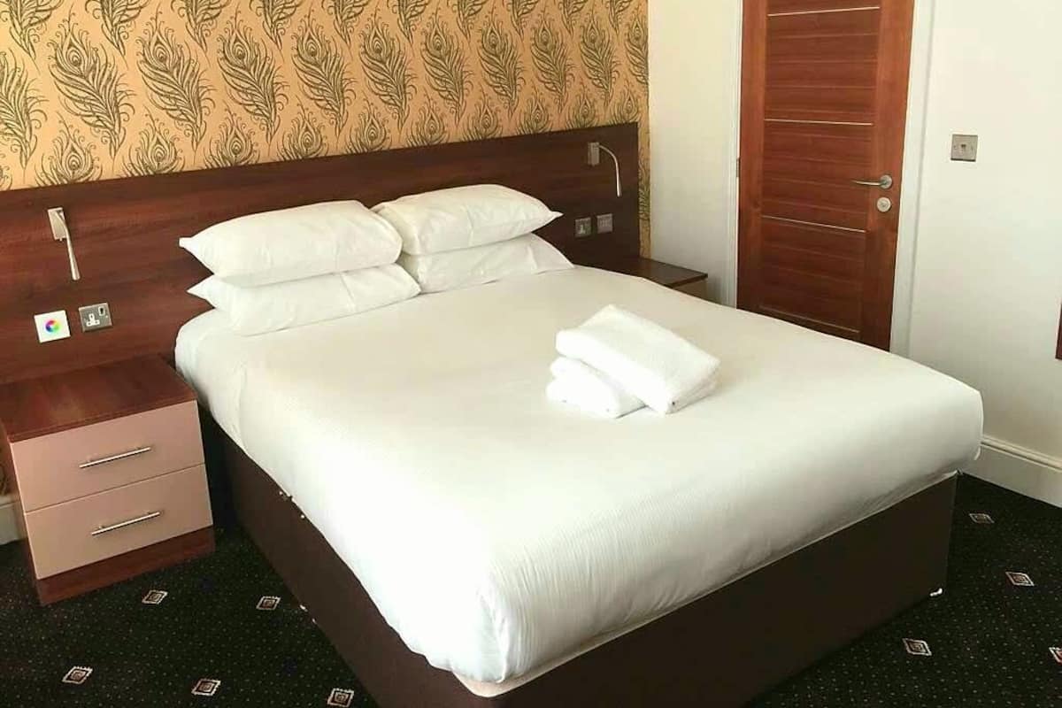 A neatly made bed with white linens and pillows, a wooden headboard, a nightstand with a drawer, and a patterned wallpaper on the wall.