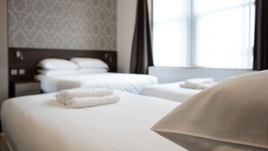 Ein Hotelzimmer mit zwei Betten, einem gemusterten Wandteppich, einem Fenster mit Vorhängen und einem ordentlich bezogenen Bett mit Kissen und Handtuch.
