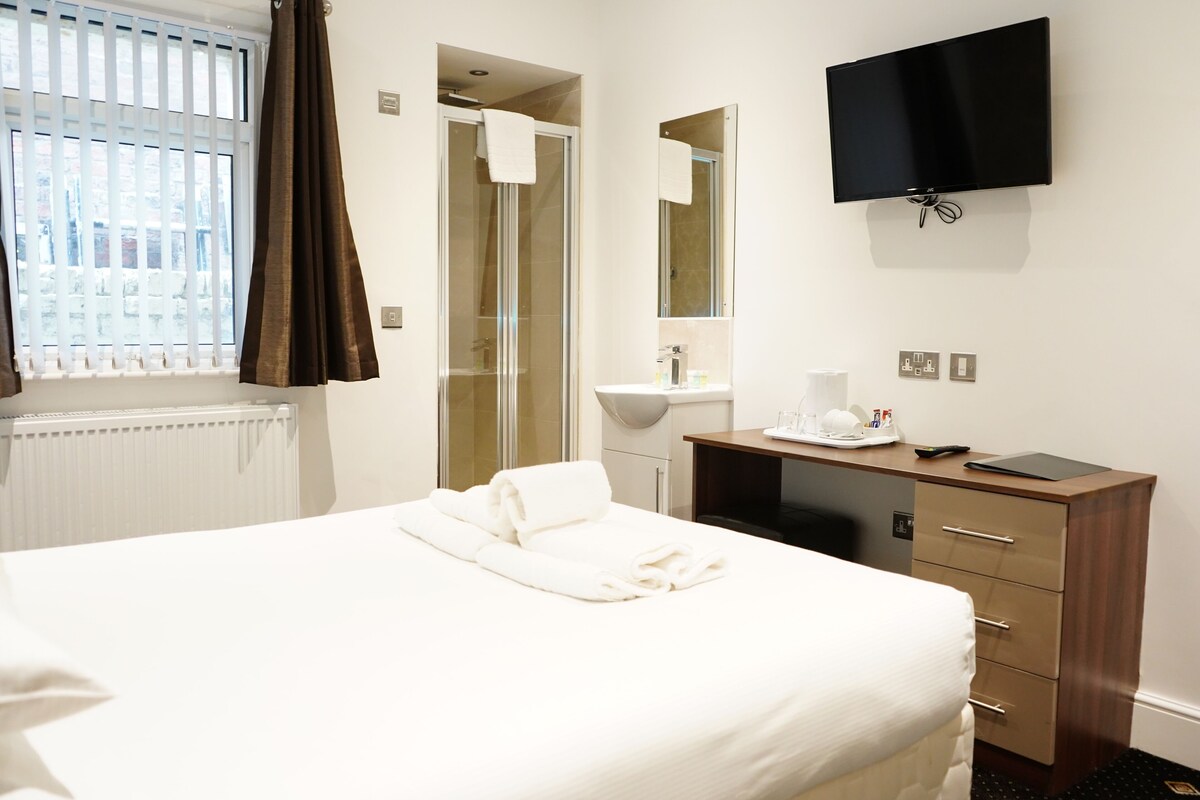 A hotel room with a bed, a desk with a computer, a TV, a bathroom with a shower, and a window with curtains.