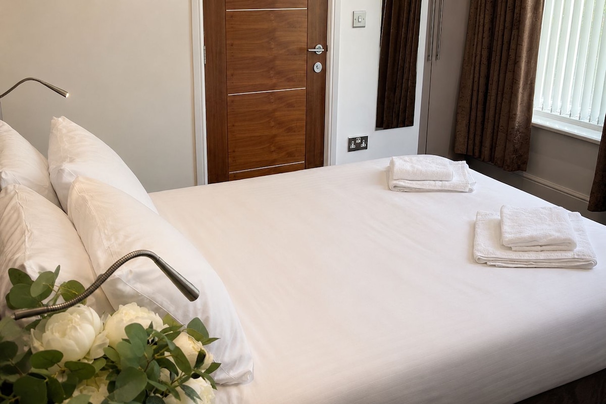 A hotel room with a bed, pillows, a bedside table, a door, and a window with curtains.