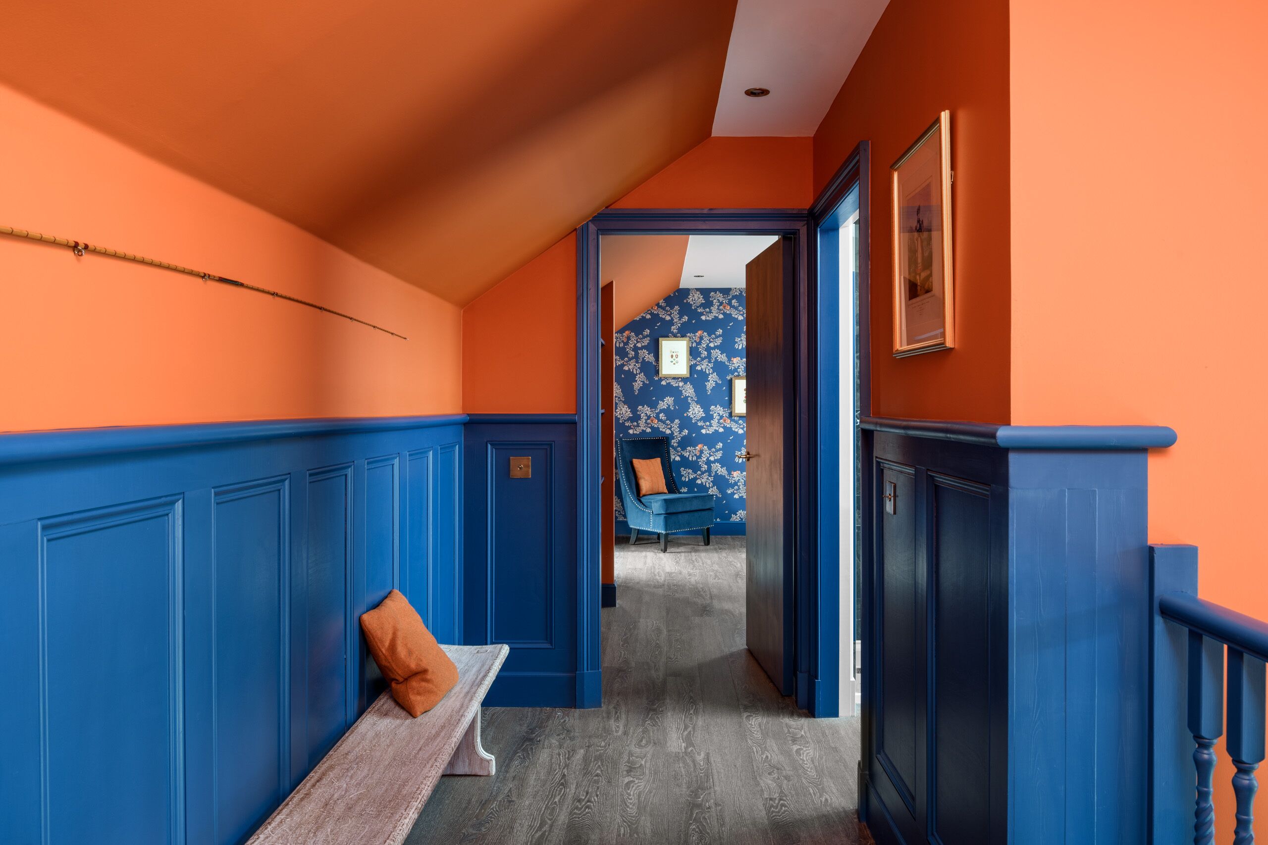 Un couloir avec des lambris bleus, un banc agrémenté d’un coussin et une porte donnant sur une pièce dont les murs sont recouverts d’un papier peint à motifs floraux bleus.