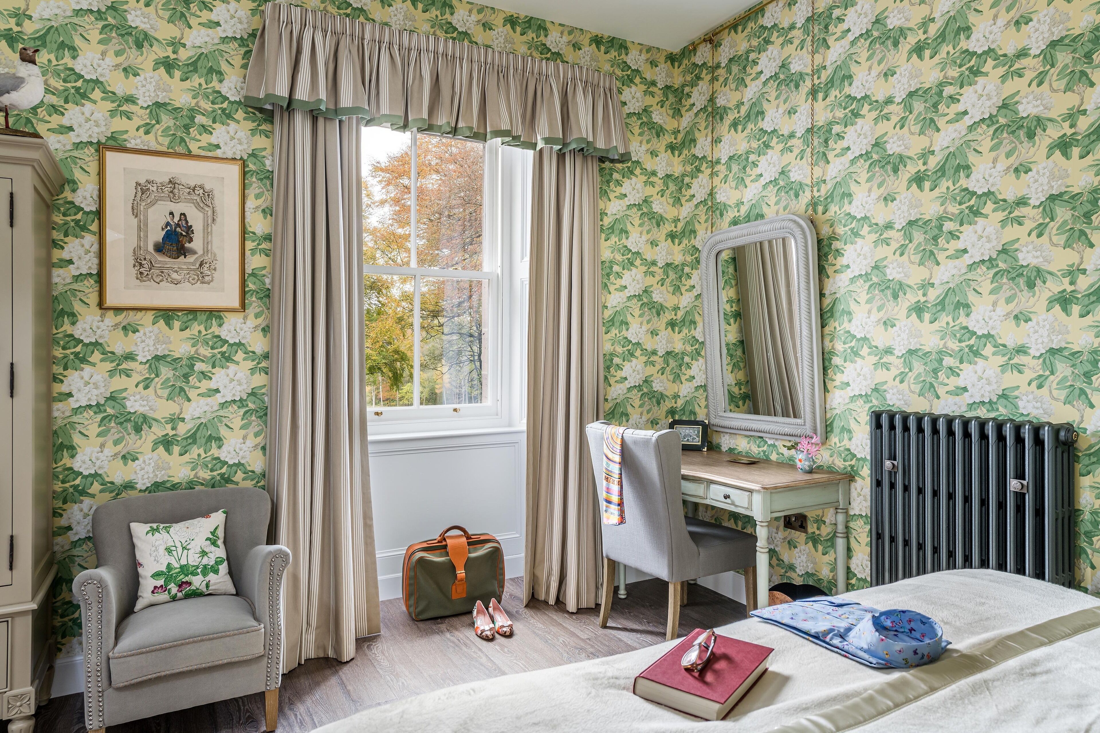 A bedroom with floral wallpaper, a window with curtains, a desk with a mirror, a chair, a radiator, and a bed with a book and a bowl.
