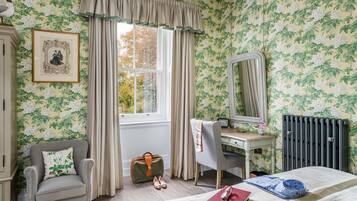 A bedroom with floral wallpaper, a window with curtains, a desk with a mirror, a chair, a radiator, and a bed with a book and a bowl.