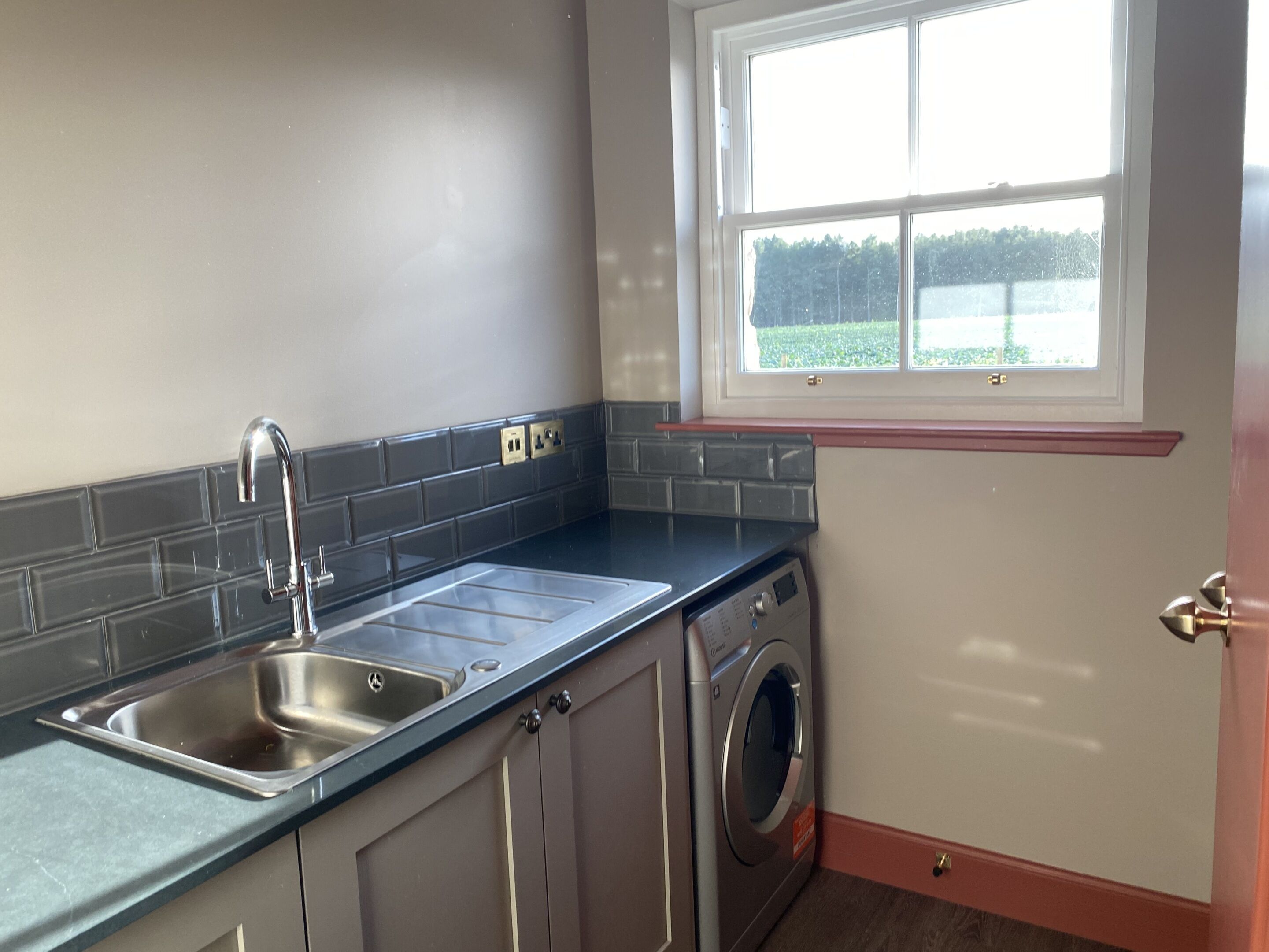 A kitchen with a stainless steel sink, a built-in washing machine, and a window with a view of greenery.