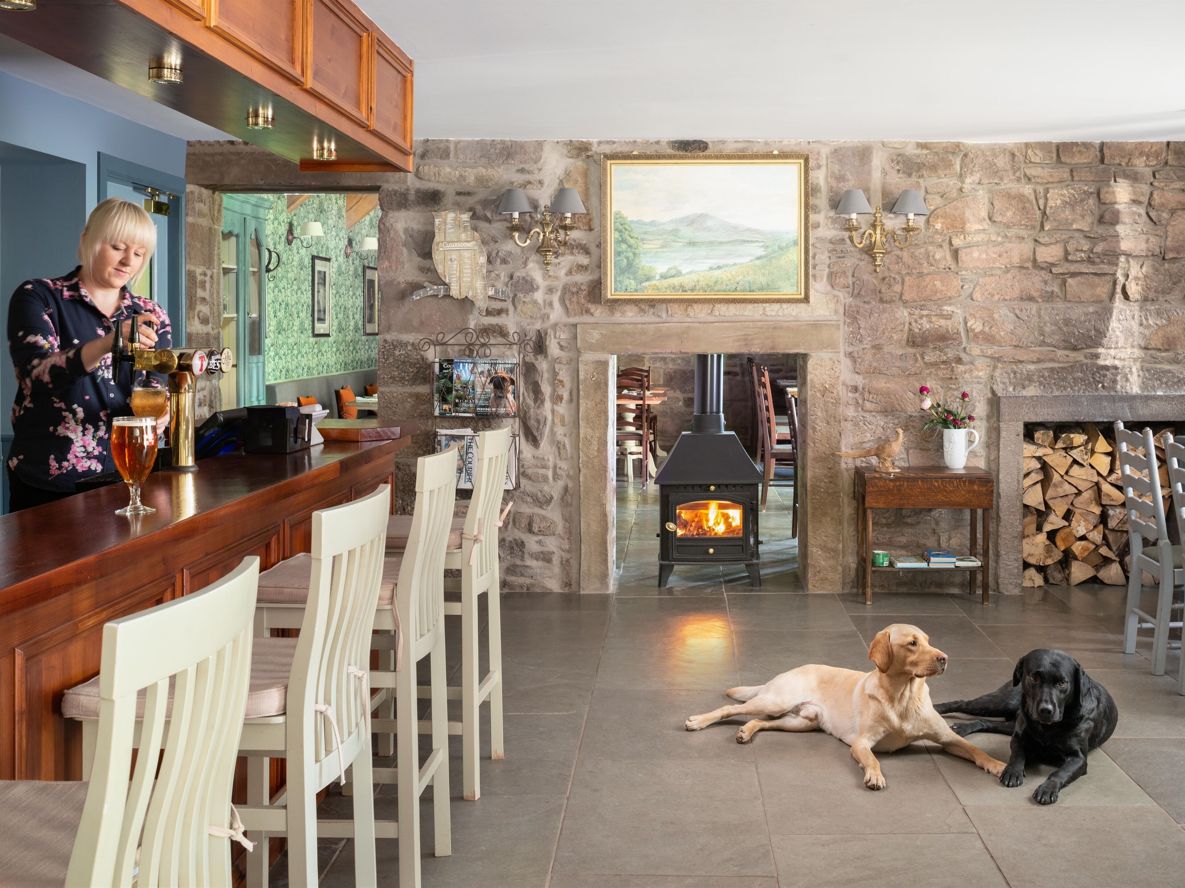 A bar area with a woman pouring a drink, a fireplace with a dog lying nearby, and a stone wall with a painting.