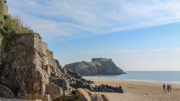 A beach with rocky cliffs, a small fort on a hill, and a few people walking on the sand. - Little Red Rooster Rest and Spa (Tenby)