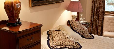 A neatly arranged bedroom with a wooden nightstand, a bed with patterned bedding, a painting on the wall, and a window with curtains.