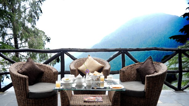 Un ensemble de mobilier de jardin en osier comprenant une table en verre et des chaises, une petite table avec un service à thé, et une vue sur une montagne.