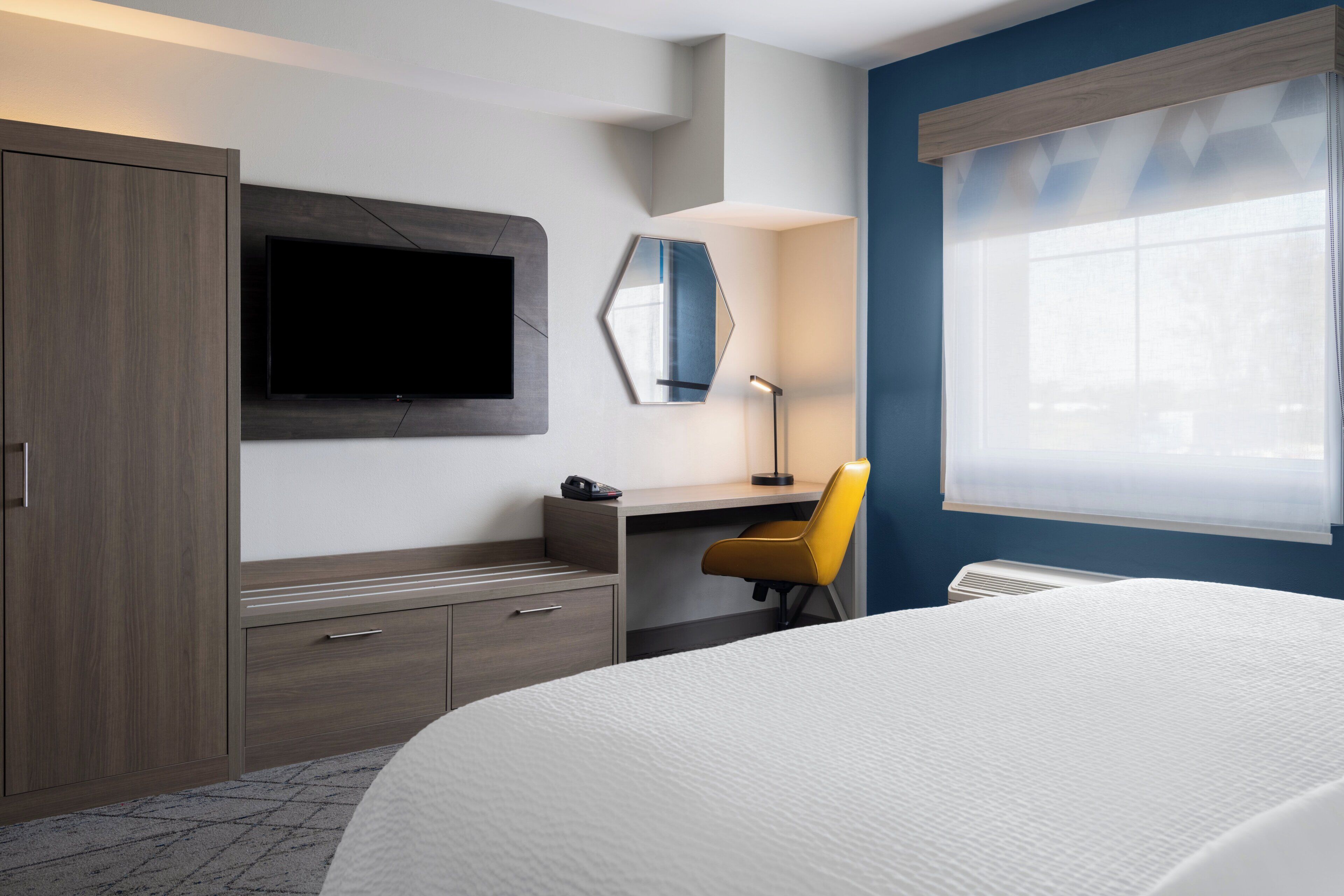 Habitación de hotel con cama, escritorio con silla, televisor montado en la pared, espejo y ventana con persianas.