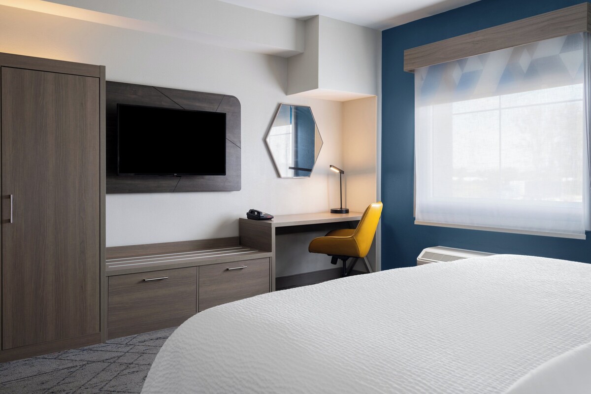 A hotel room with a bed, a desk with a chair, a TV mounted on the wall, a mirror, and a window with blinds.