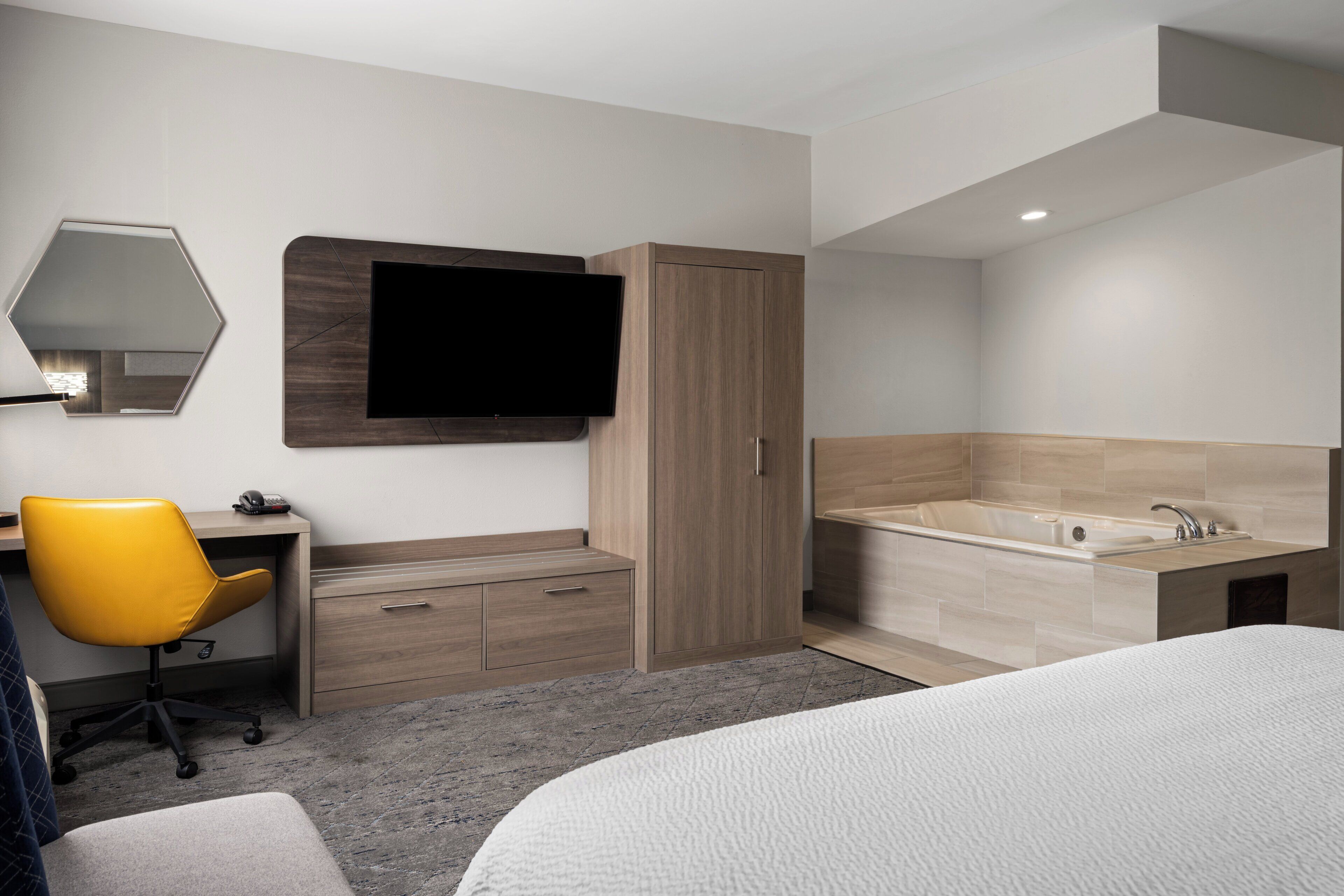 A modern hotel room with a bed, a desk with a chair, a large mirror, a flat-screen TV, a wooden wardrobe, and a bathtub.