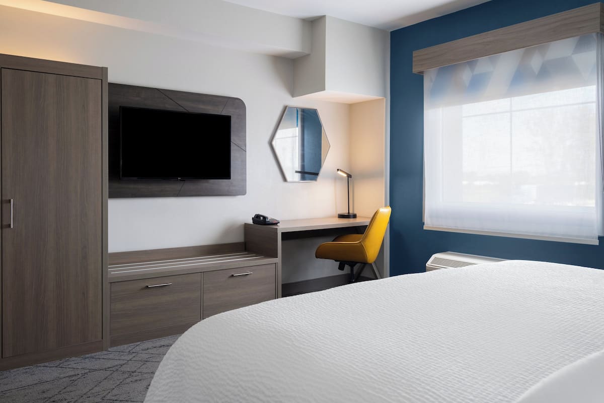 A hotel room with a bed, a desk with a chair, a TV mounted on the wall, a mirror, and a window with blinds.